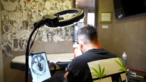 rear shot of a tattoo artist working on a client while referencing a dog image on a tablet in the studio