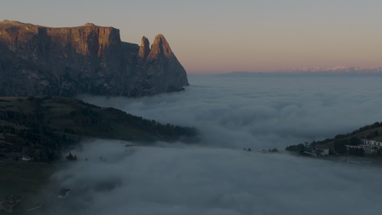 Cinematic aerial view of frosty Alpe di Siusi meadows and scattered mountain huts at dawn, mist drifting across rolling hills beneath the dramatic Sassolungo peaks in the Dolomites, Italian Alps