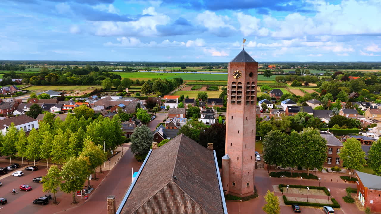 Vista aérea de una iglesia de ladrillo con una alta torre de reloj. Una destacada iglesia de ladrillo con una alta torre de reloj cuadrada situada en una plaza del pueblo con un aparcamiento y árboles verdes.
