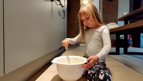 Cute-smiling-caucasian-girl-sitting-on-floor-and-eating-leftover-dough-from-bowl-after-baking-vanilla-muffin