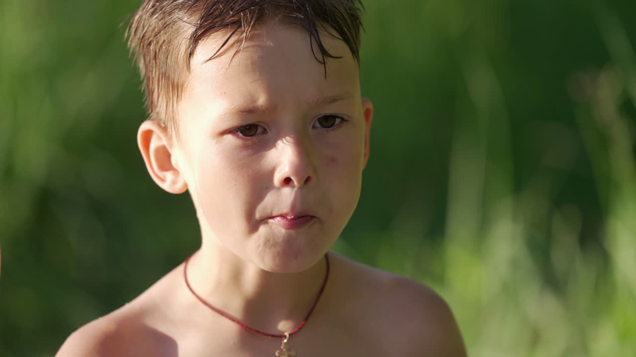 Portrait of a cute hungry boy outdoors. Little boy with wet hair is eating something on blurred background in summer.
