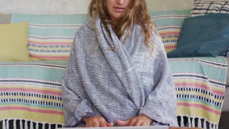 Thoughtful-caucasian-woman-sitting-on-floor-wearing-blanket,-using-laptop-in-cottage-living-room