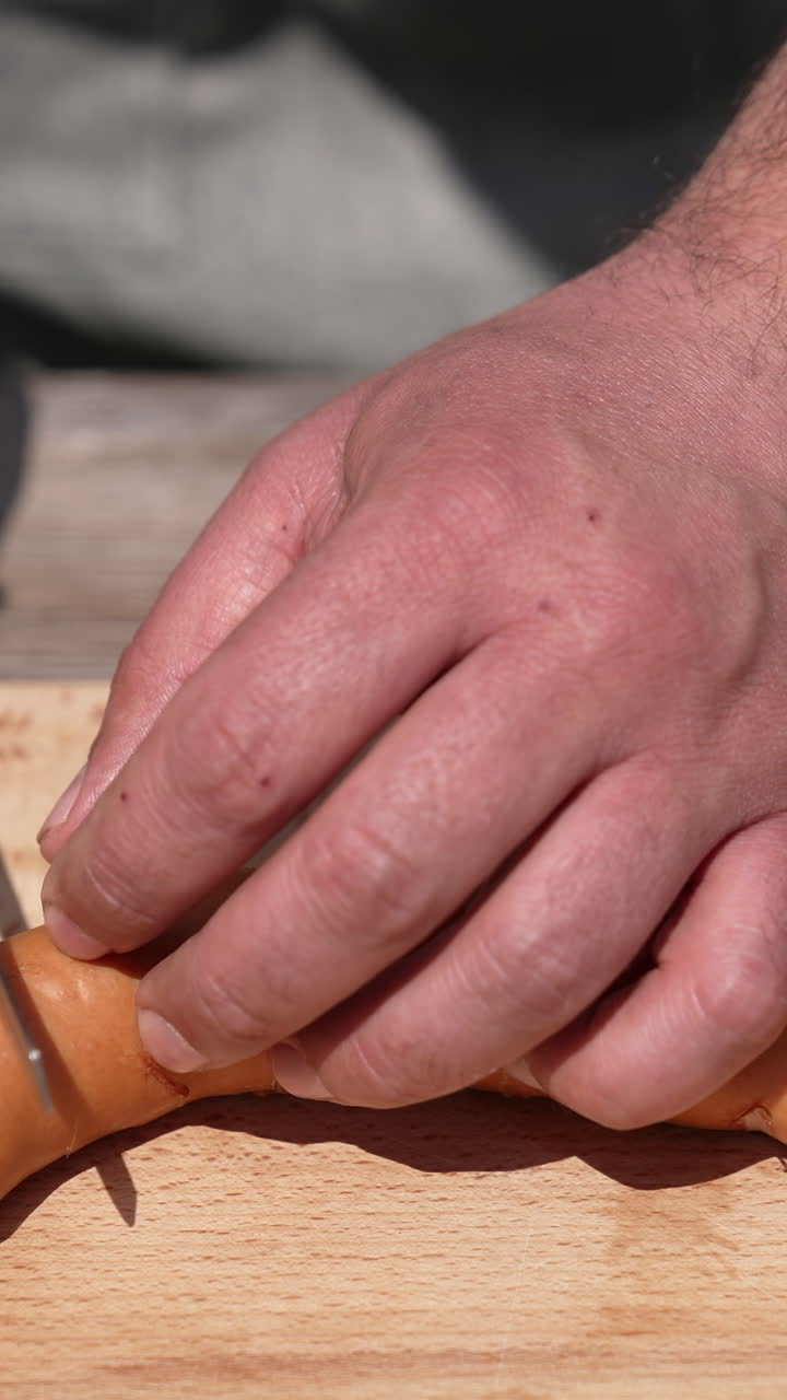 Man's hands prepare sausages for barbeque. Chef in doing incisions on juicy sausages with a knife on a wooden board before grilling the outdoors. Close-up. Vertical video