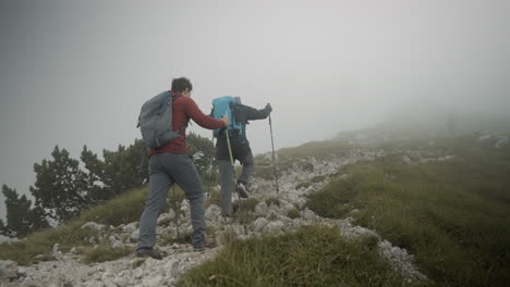 dos excursionistas caminando por una colina en un camino rocoso hacia la cima más densa de nubes de la montaña