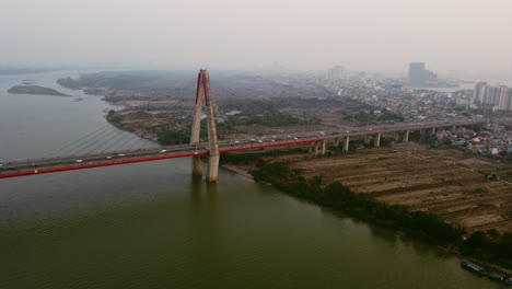 Vista-Aérea-De-Drones-Rodeando-Un-Gran-Puente-Colgante-Sobre-Un-Río-Con-La-Ciudad-Al-Fondo-En-Hanoi-Vietnam