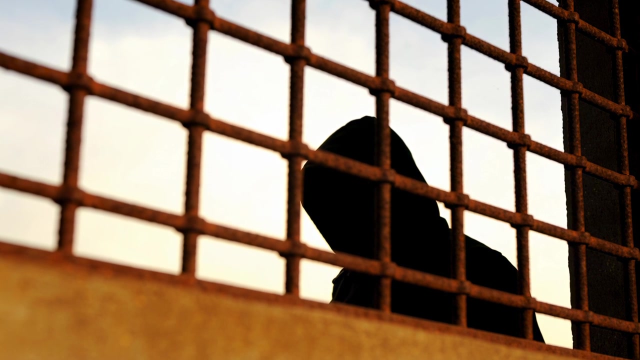 Leaning hooded person shifting forward behind rusted grid on concrete ledge, warm sky backlighting