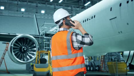 vista trasera de un ingeniero masculino asiático con casco de seguridad hablando en teléfono inteligente mientras está de pie con el avión en el hangar