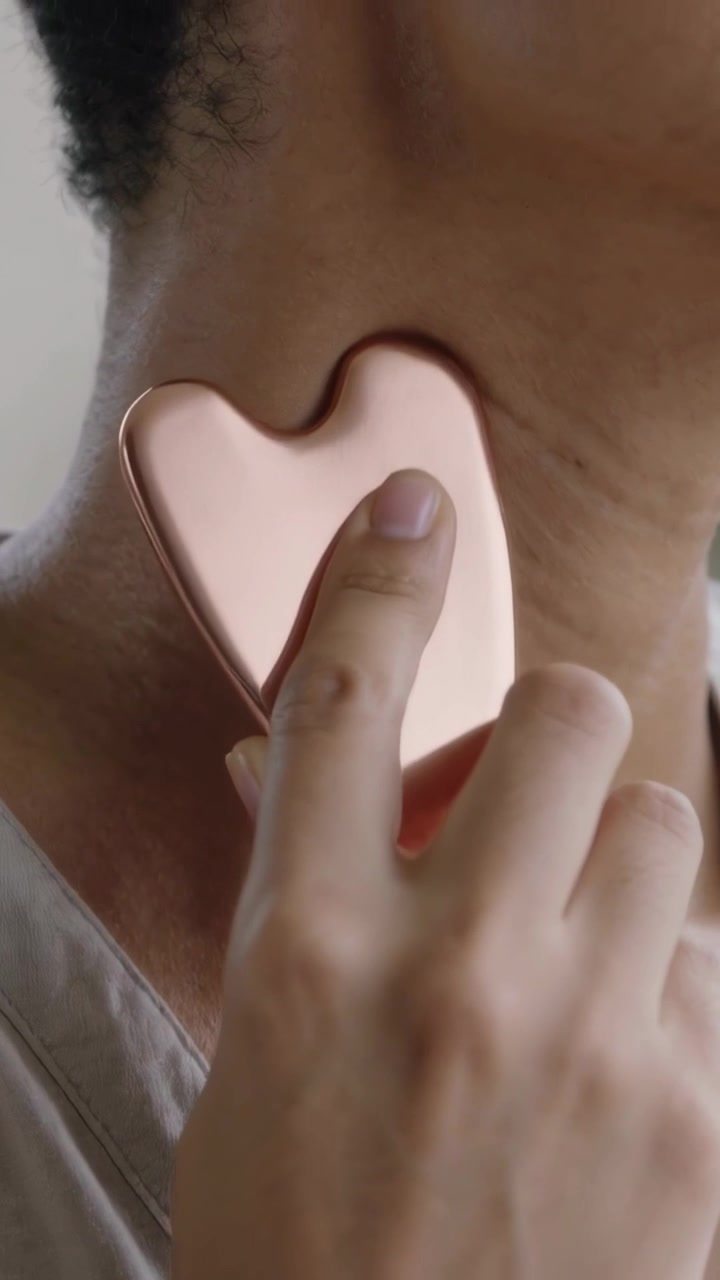 Video verticale: Posizionando un gua-sha di quarzo rosa, un uomo si massaggia il collo con la mano destra, indossando una maglietta con scollo a V, a casa.
