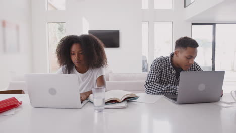 Black-teenage-boy-helping-his-sister-while-they-sit-at-home-using-laptop-computers,-front-view
