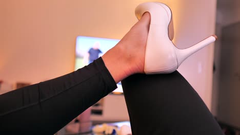 una mujer con polainas negras se sienta en el sofá y ve la televisión mientras mueve sus tacones blancos de cuello alto de un lado a otro