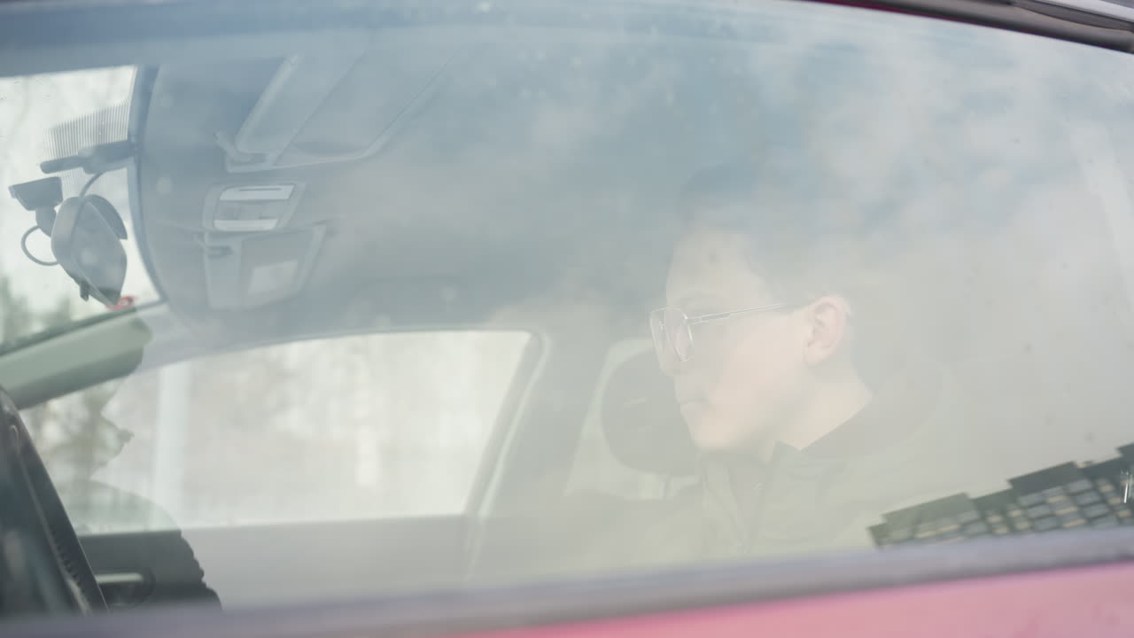 side view of man in sunglasses and winter jacket seated inside vehicle rolling up window with neutral expression while snow-covered scenery reflects on glass under soft daylight