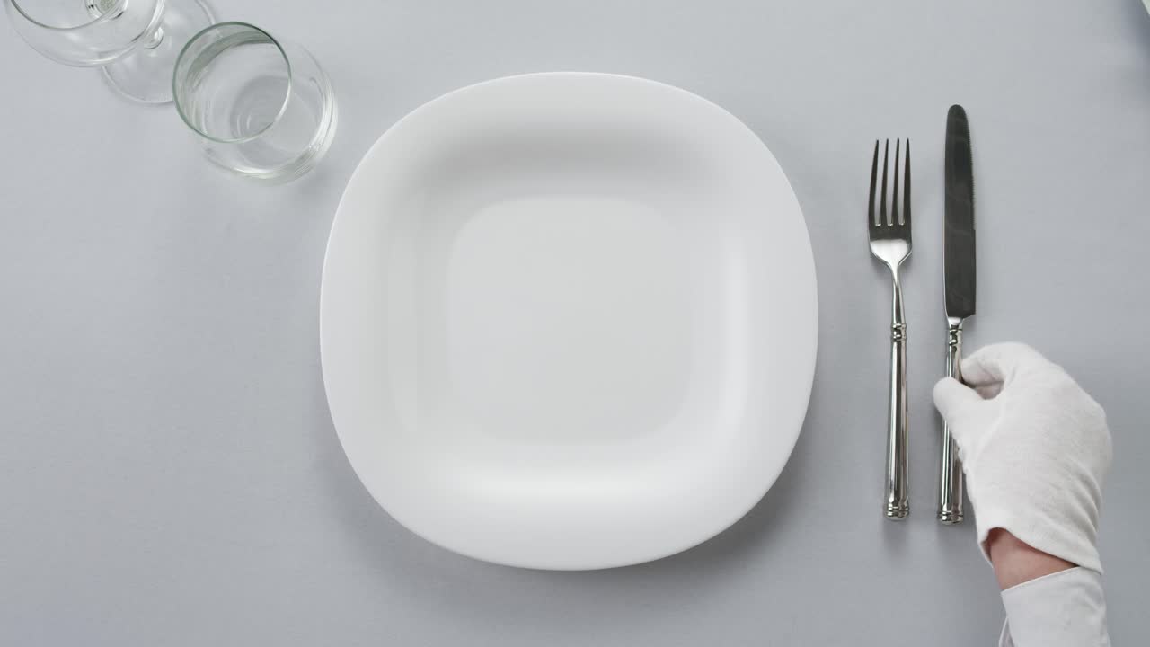 Professional table setting with white gloves. Plates, fork, knife, and glass arranged on a white table.
