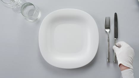 professional table setting with white gloves. plates, fork, knife, and glass arranged on a white table.
