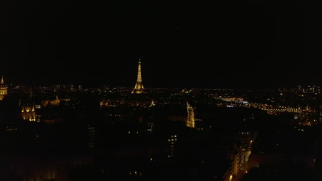 Fly-above-large-city-at-night.-Illuminated-streets-and-landmarks.-Famous-Eiffel-tower-glowing-into-darkness.-Paris,-France