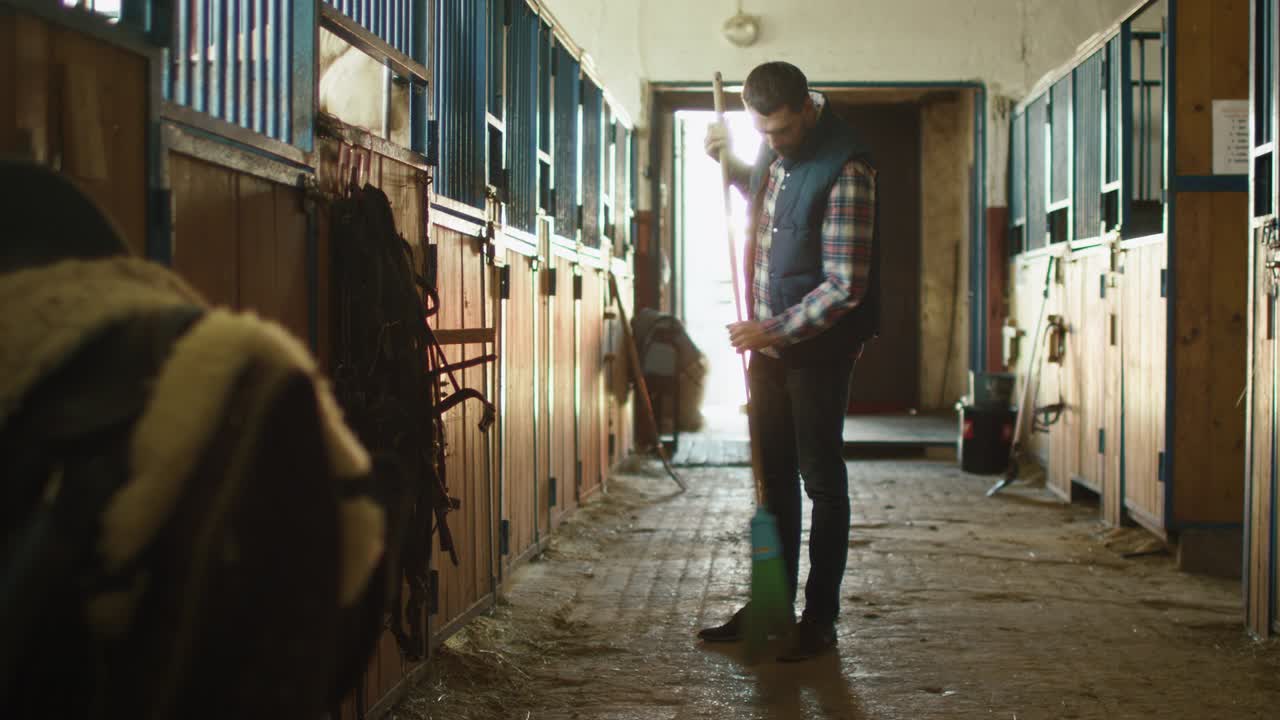 Man is cleaning a stable with a broom on a sunny day.