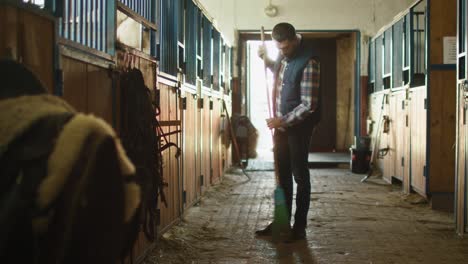 man is cleaning a stable with a broom on a sunny day.