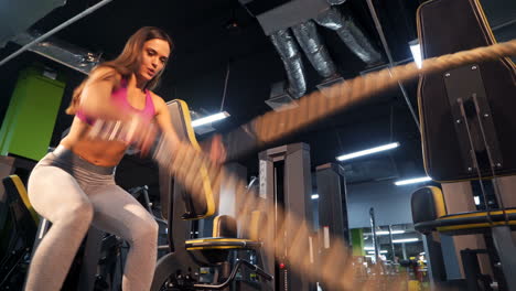 strong girl with long dark hair trains triceps by shaking cables on the background of sports equipment. happy young woman in sportswear doing workout inside. close-up