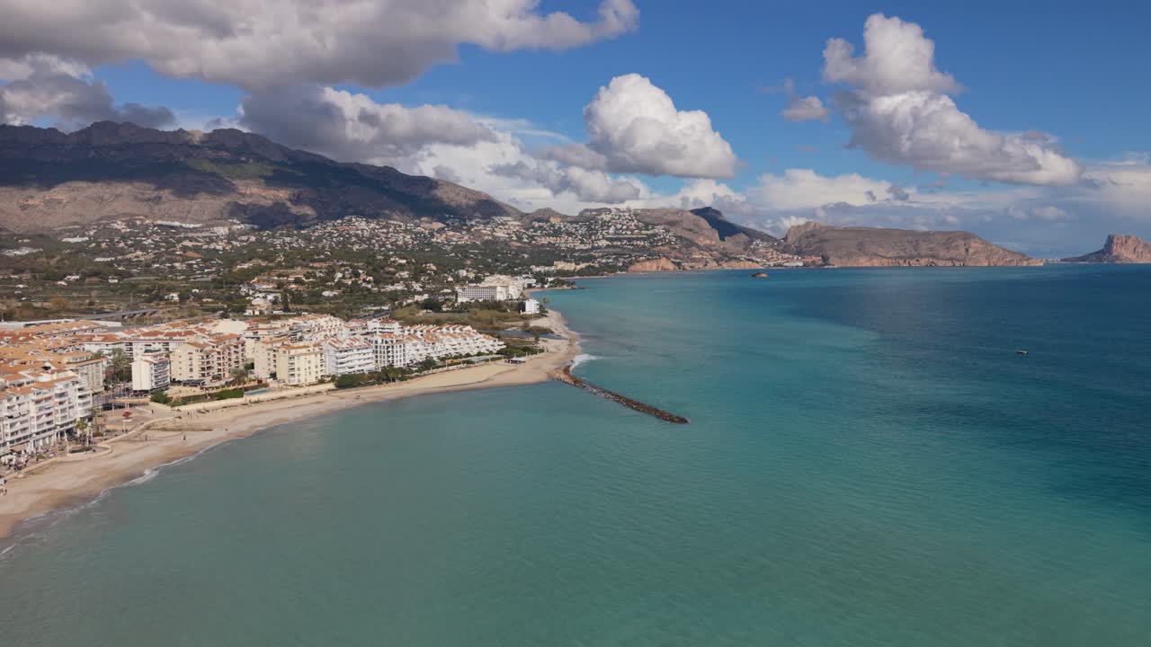 Albir e Altea Costa Blanca Spagna. Volo di drone ad alta quota all'indietro sopra il mare blu cristallino della spiaggia e del frangiflutti con vista sull'Isola del Pavone, le montagne e nuvole interessanti sotto il sole splendente