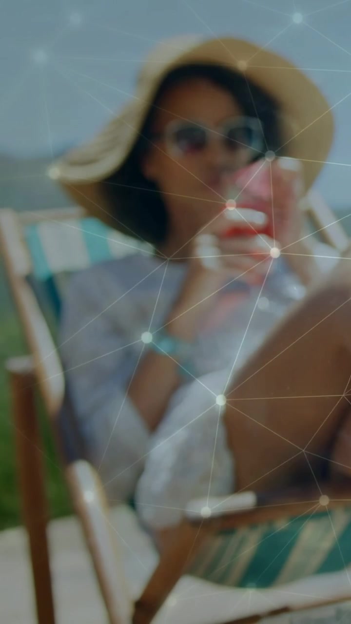Vídeo vertical: Mulher em uma cadeira levantando uma bebida rosa para lazer, enquanto uma sobreposição de rede brilha no colo.