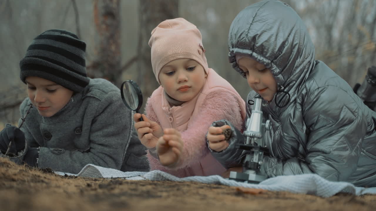 Little girls and boy looking through a magnifying glasss and microscope in the forest. Kids interested in nature and science. Close-up.