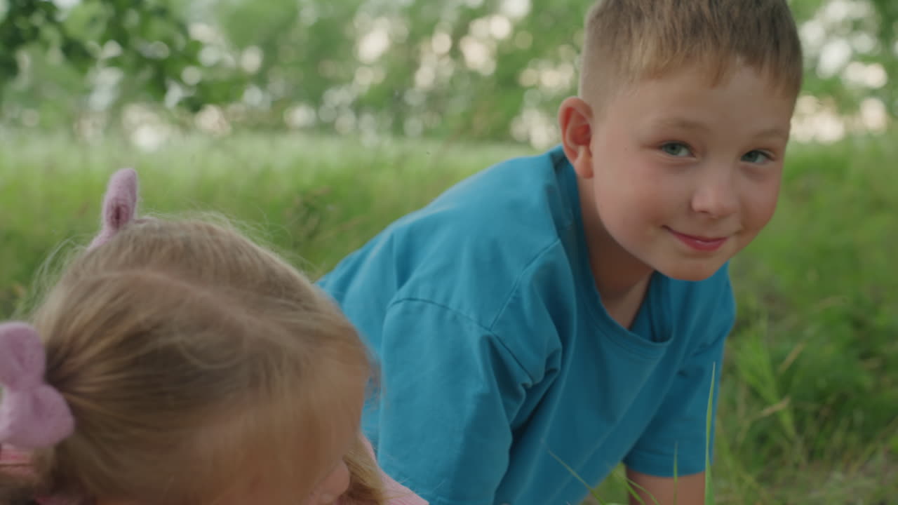 수박을 안고 있는 어린 백인 소녀, 뒤에 미소를 짓는 남자아이, 풀과 나무가 있는 여름 초원에서의 피크닉, 따뜻한 햇살, 어린 시절의 순수함과 형제자매 간의 유대감을 담은 솔직하고 장난기 넘치는 순간