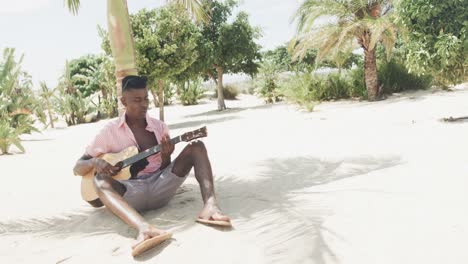 Feliz-Hombre-Afroamericano-Sentado-Bajo-Un-árbol-Tocando-La-Guitarra-En-Una-Playa-Soleada,-Cámara-Lenta