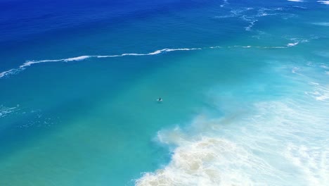 Surfer-alone-in-turquoise-ocean-waiting-on-waves,-high-angle-drone-shot