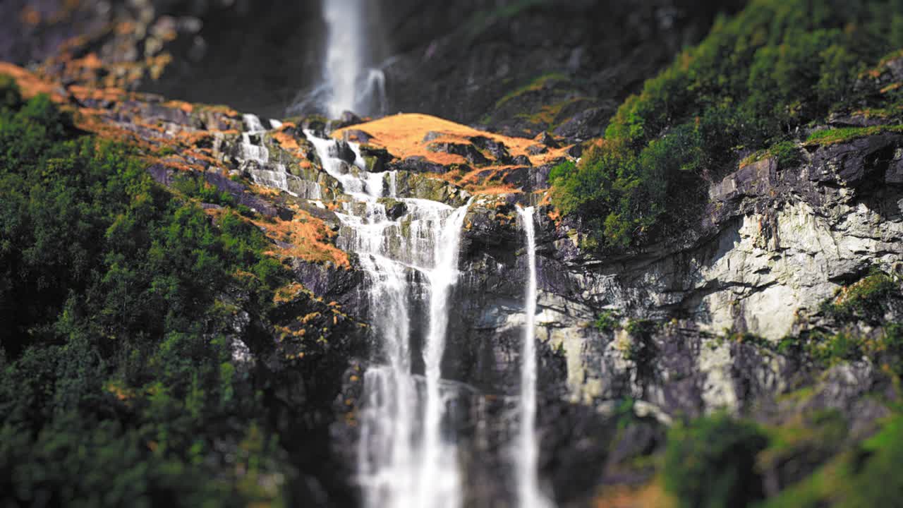 una vista en miniatura de un primer plano de una cascada de dos niveles en los acantilados cubiertos de bosque por encima del lago loenvatnet