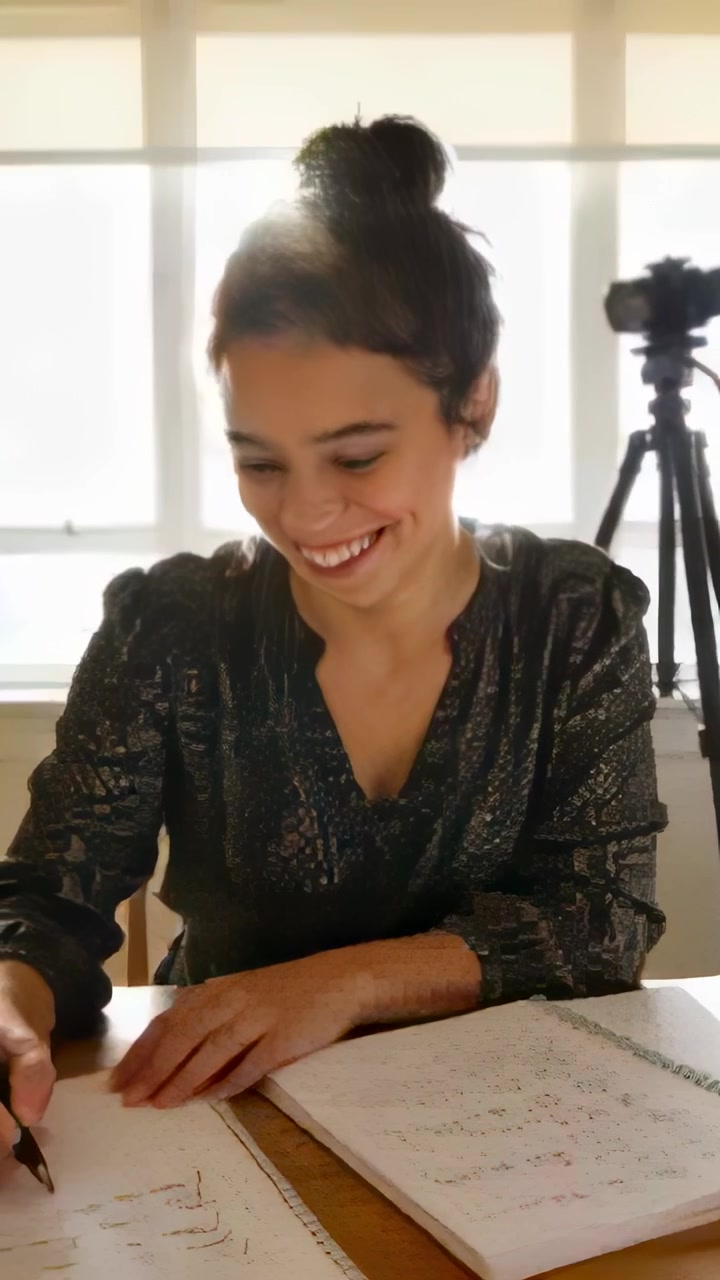 Video vertical: Mujer con blusa escribiendo en la mesa de casa, sonriendo a una señal fuera de cámara y tomando notas.