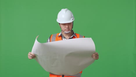 asian male engineer with safety helmet looking at blueprint in his hands and looking around while standing in the green screen background studio