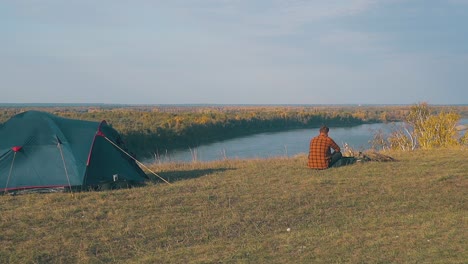 guy-in-orange-shirt-sits-at-bonfire-by-blue-tent-in-morning