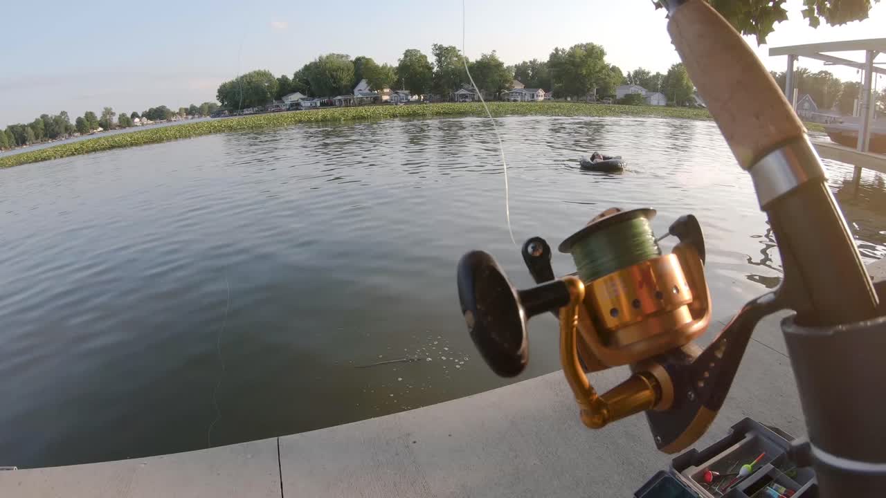 UHD, slow motion shot of a fishing pole with a person relaxing in a lake on an inter tube in the background during the summer.