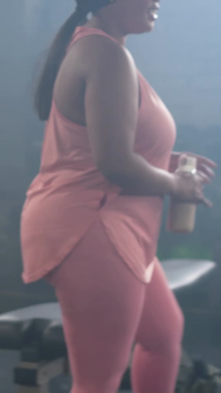 Video verticale: Entrando da sinistra, donna in rosa che posa lo shaker vicino alla borsa, legandosi i capelli per l'allenamento in palestra.