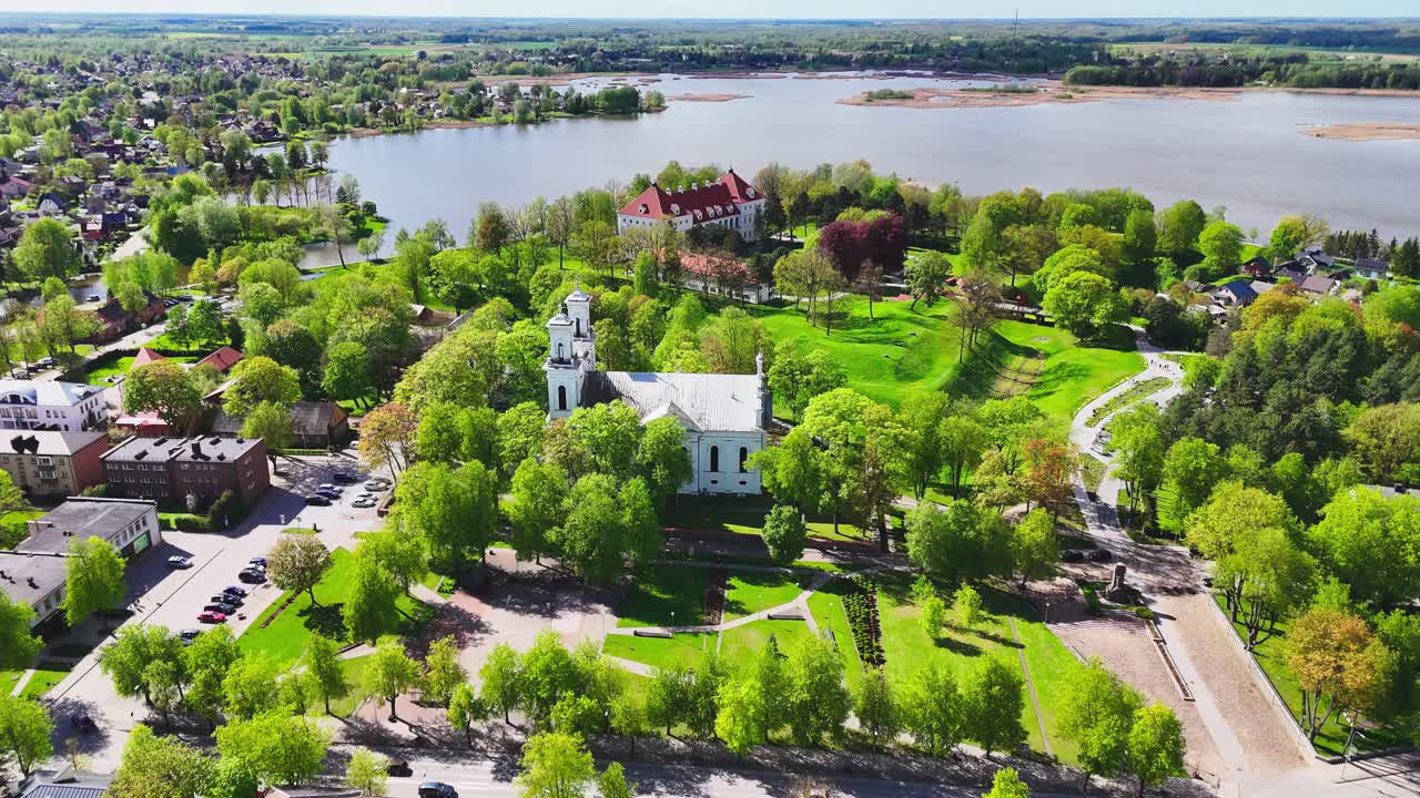 A white church and red-roofed estate rise among lush green trees near a lakeside in a scenic European town surrounded by spring colors and distant rooftops.
