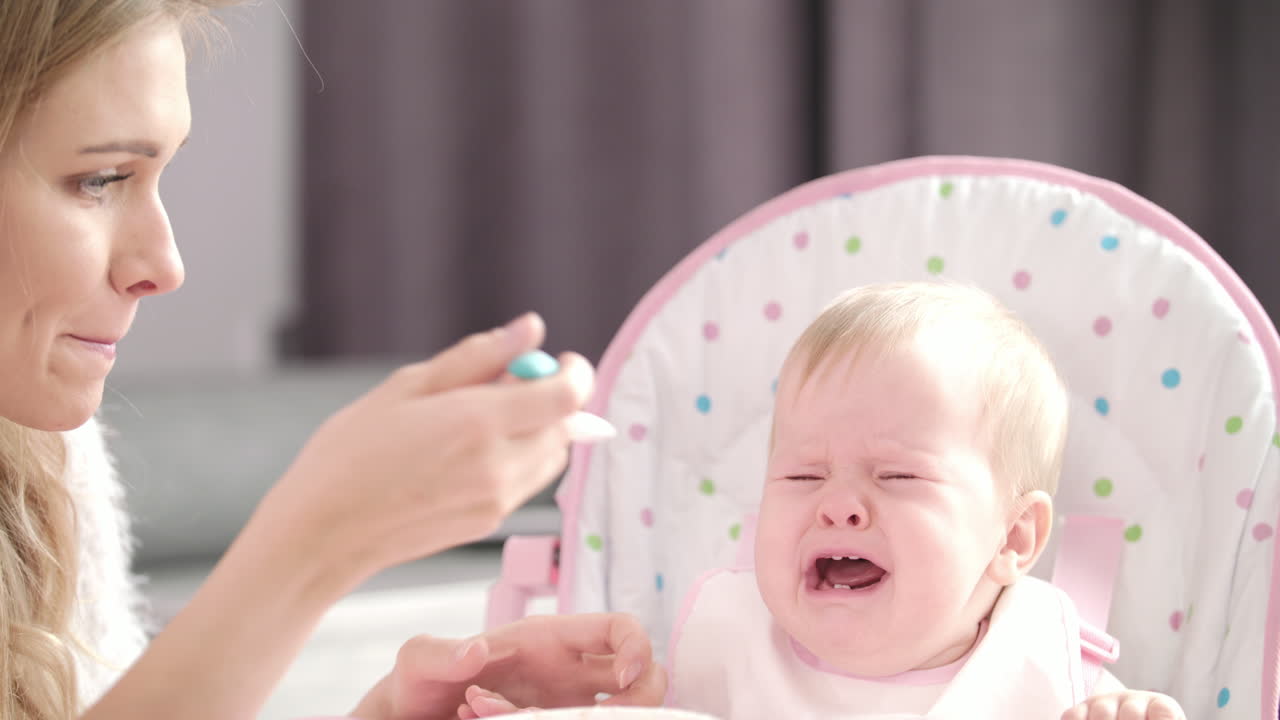 Mom feeding baby with spoon. Little kid crying in baby chair