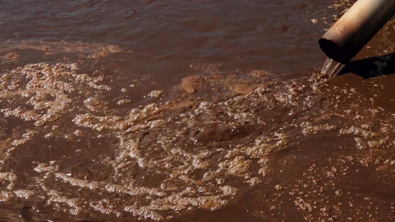 Rusty pipe discharging muddy brown wastewater into a polluted stream, with foamy surface and visible bubbles