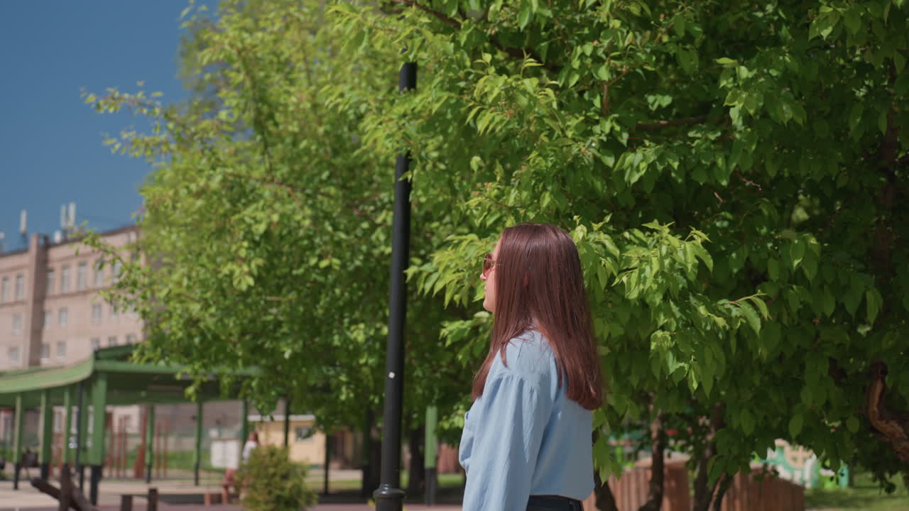 햇빛과 그늘 아래에서 나무를 관찰하는 여성, 현대적인 건물 아래에서 잎이 무성한 주변을 살펴보는 여성, 햇빛이 쏟아지는 도시 공원 환경에서 데님 셔츠를 입은 백인 여성