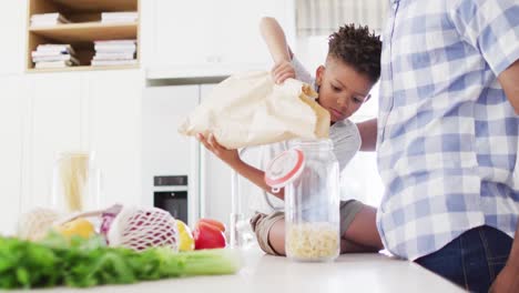 African-american-father-and-son-filling-storage-jar-with-pasta-in-kitchen,-slow-motion