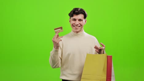 el hombre está mirando a la cámara y sonriendo. está sosteniendo bolsas de compras y agitando una tarjeta de crédito. está de pie sobre un fondo verde. pantalla verde. 4k.