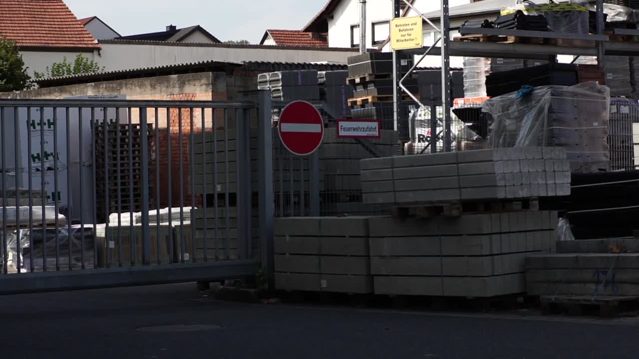 A view into an outdoors warehouse yard behind a metal fence, showcasing various construction materials stacked on multi-level racks. Ideal for themes of storage, logistics, and industrial supply.