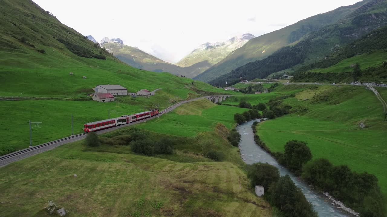 tren pasando por los pintorescos alpes suizos con pequeño puente y río al lado de las vías durante el verano en el paso de furka, filmado desde un dron en 4k
