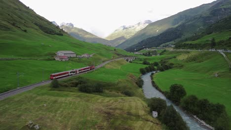 train going through the scenic swiss alps with small bridge and river next to the tracks during summer at furka pass, shot from a drone in 4k