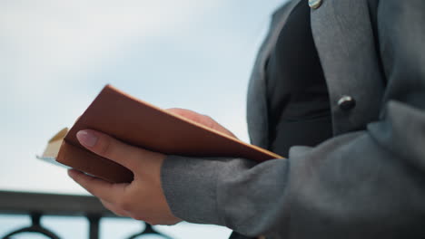 closeup of a lady holding an open book in her right hand, with her left hand supporting the book from beneath, she stands close to an iron fence, with the pages slightly lifting as if from a breeze