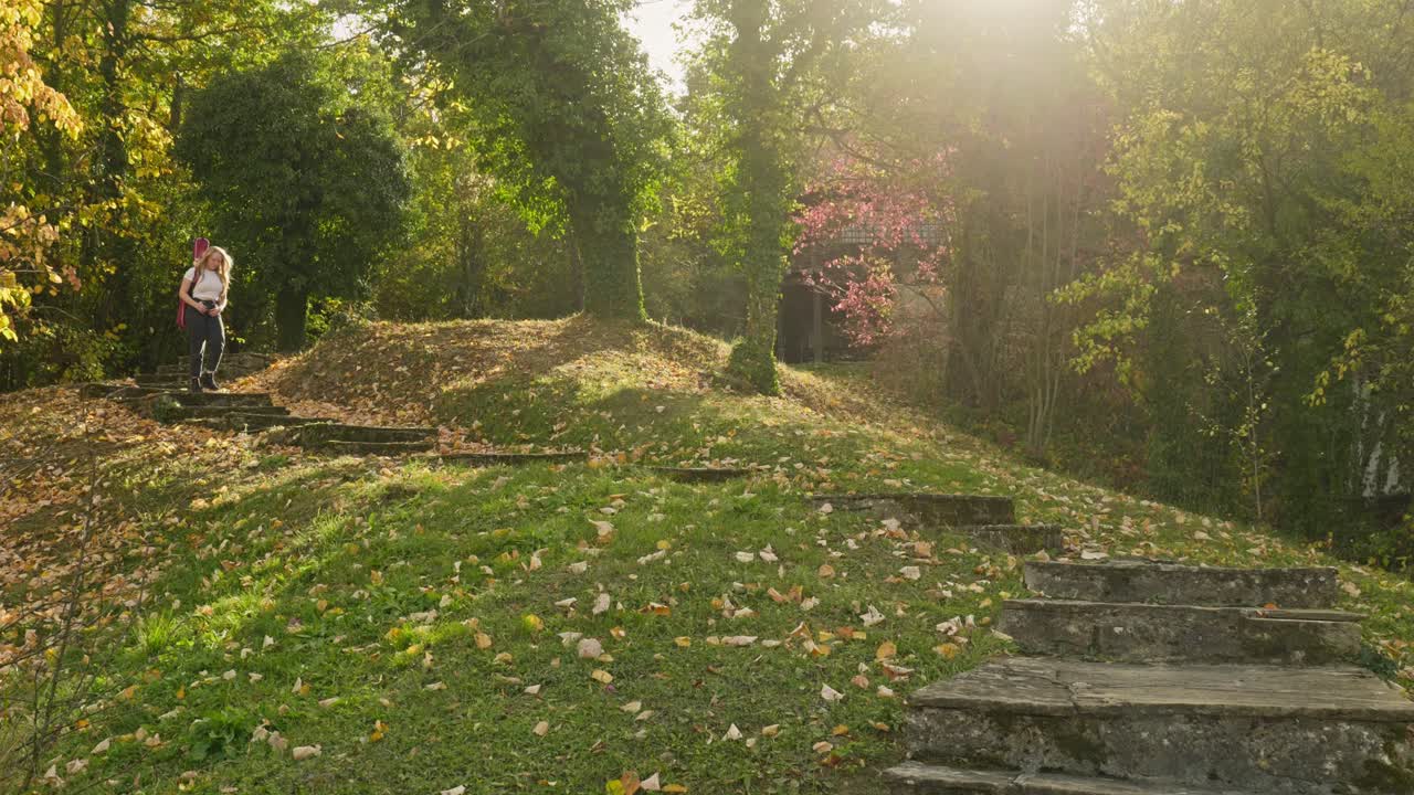 una mujer rubia músico desciende las escaleras del bosque en una escena de otoño iluminada por el sol