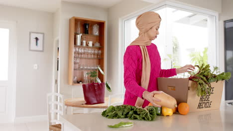 Happy-biracial-woman-in-hijab-with-grocery-shopping-in-kitchen-at-home-with-copy-space,-slow-motion