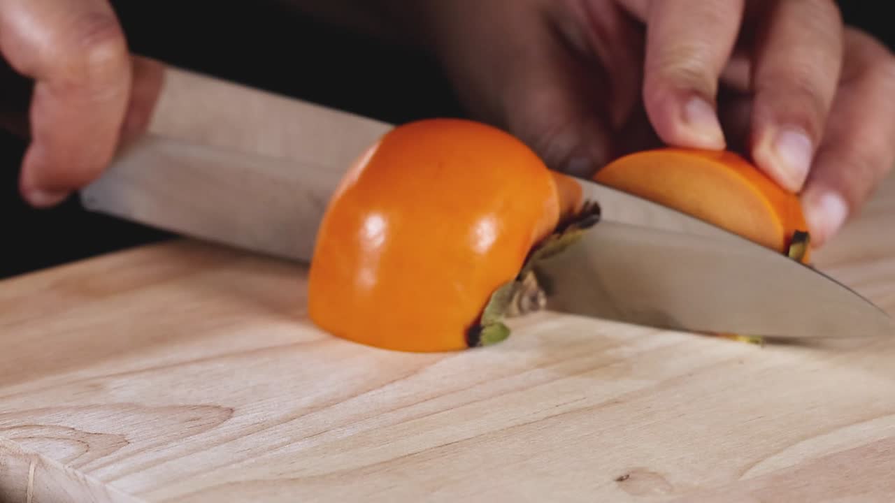 Et detaljeret billede af hænder, der skærer en halv persimmon på et træbræt med en skarp kniv