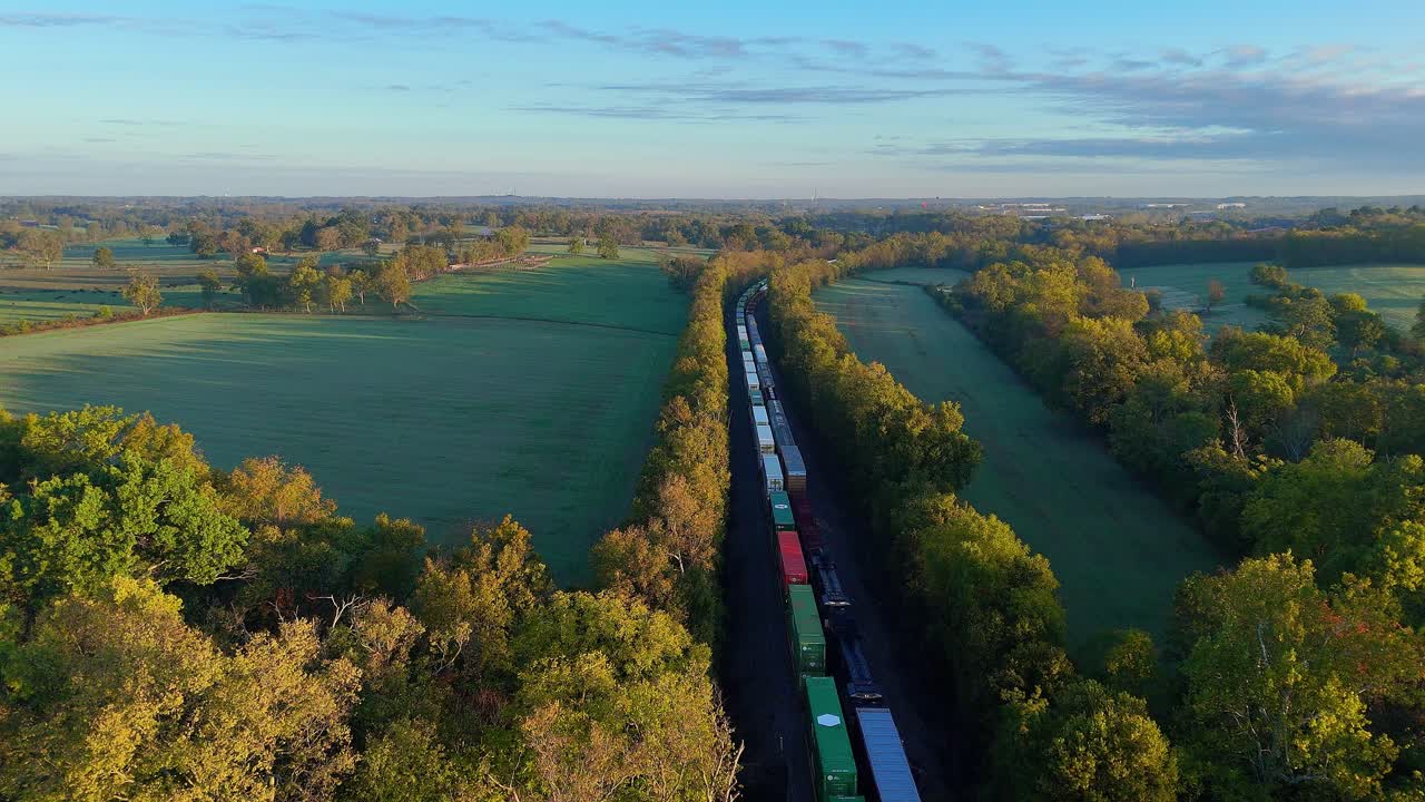 Treno merci che attraversa vasti campi verdi e alberi autunnali all'alba. Lungo treno merci che viaggia attraverso un paesaggio esteso, rappresentando il trasporto e il commercio
