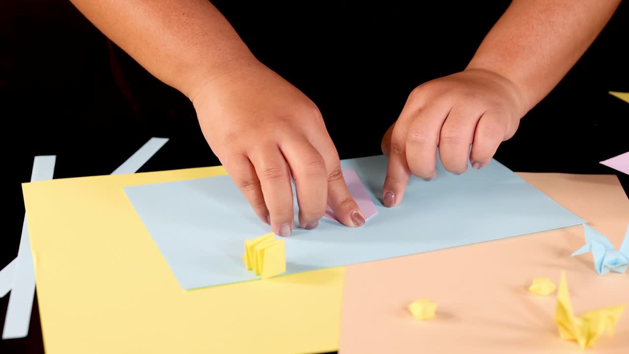 Child’s hands fold yellow origami on pastel paper, brightly lit, viewed from above