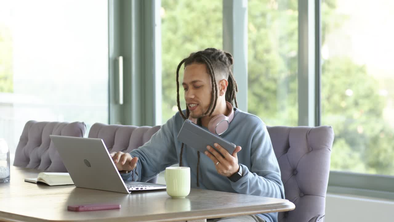 En afroamerikansk mand nipper til kaffe og skriver på sin bærbare computer, mens han bruger en tablet til at gennemgå en rapport