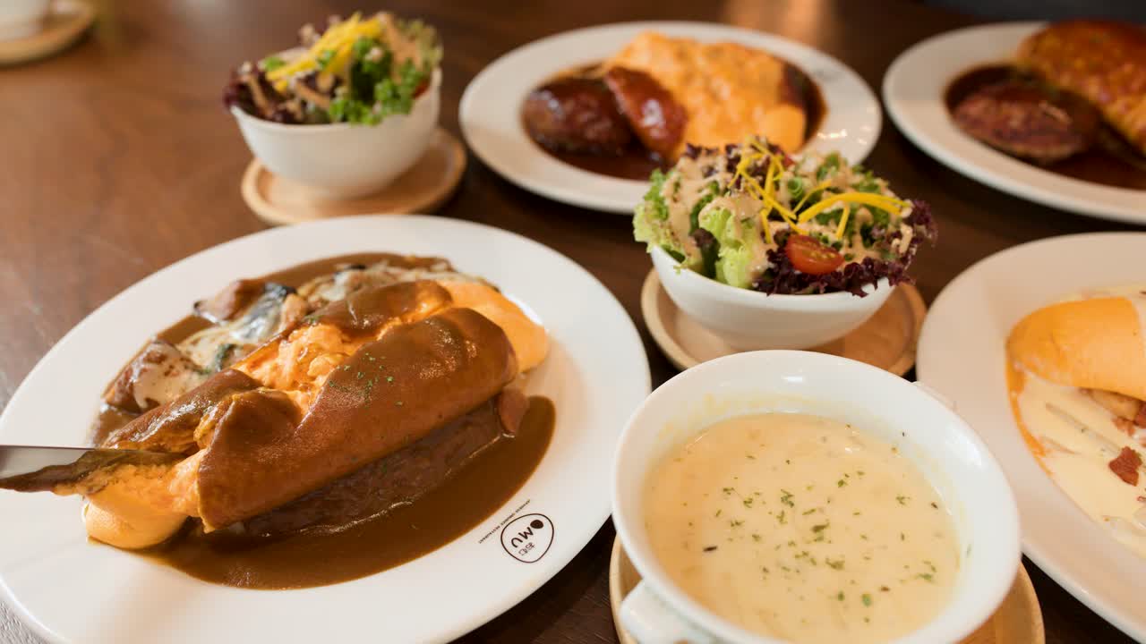 Hand slices omelette rice with creamy sauce, soup, salad, and bread in bright restaurant setting